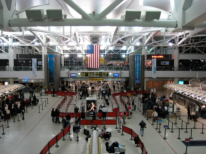 Crowded check-in hall under white steel trusses, with a large flag hanging