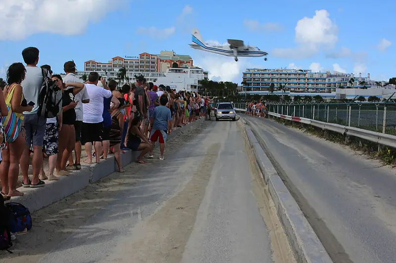 Crowd lined along a fence beside a sandy road as a jet flies low overhead