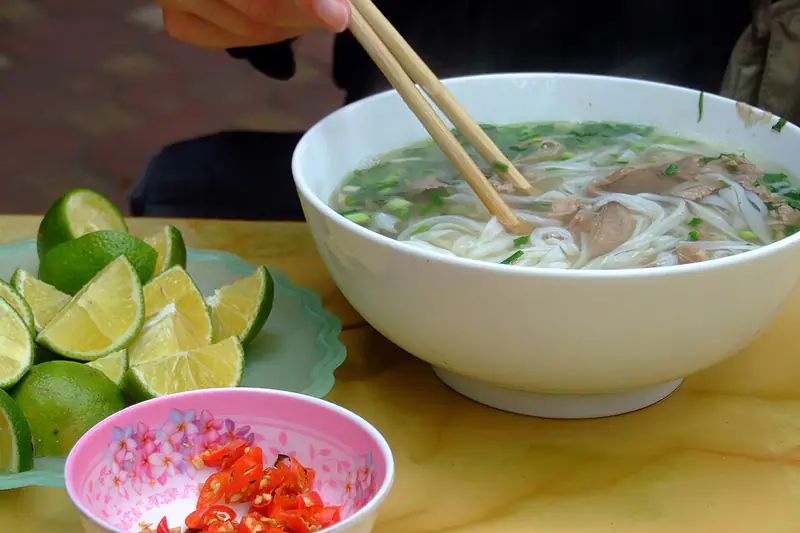 Bowl of clear noodle soup with sliced meat and herbs, with limes and chilli beside it